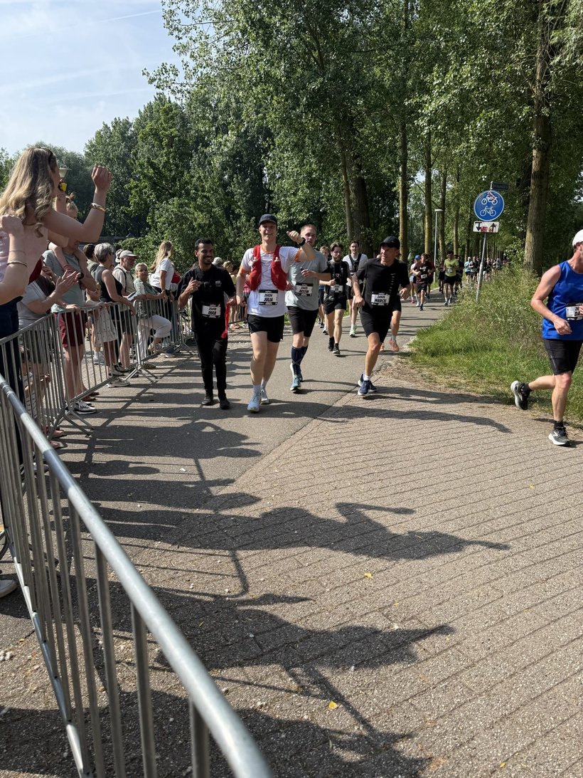 Runners in a race on a sunny day on a cycle path between tall trees.
In the middle, a man is running wearing a white shirt, black shorts, a red running vest and a black cap; he is smiling and has a race number on his stomach.
Around him, several other participants are running, mostly in black or dark sportswear, with race numbers on their stomachs.
On the left, behind barriers, there is a row of spectators standing close together; some are clapping, others are holding up a phone to film or take photos.
On the right, there is a strip of grass along the path and further away more runners in the distance.
Above the path, there is a blue circular traffic sign with a white bicycle symbol indicating that it is a cycle path.
The sun is shining brightly, casting long shadows of the spectators and the barriers on the asphalt.