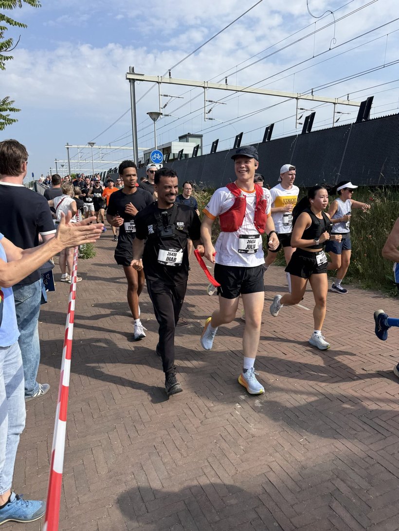 Group of runners during a race on a brick path alongside a railway line.
At the front, two men are running next to each other. On the left, a man with a medium skin tone in completely black sportswear, including black trousers, T‑shirt and shoes. He is wearing a race bib on his chest with the text “26.49 IHAB” and is smiling broadly.
To his right, a man with a light skin tone in a white T‑shirt, black shorts, white socks and white running shoes with yellow soles. He is wearing a red running vest and a white cap; he is also smiling. On his chest hangs a race bib with “26.49 JOSUA”. Between the two there is a red strap or cord that connects them, as if they are running as a duo.

Behind and beside them, several other participants are running in sportswear in different colours, including black, white and orange. On the left side of the course, spectators are standing close to a red‑and‑white tape; a few hands are stretched out in an encouraging clapping gesture.

On the right side of the path there is tall grass, and on the left an elevated railway line with overhead wires and a fence. Further back, a blue road sign with a bicycle on it can be recognised, indicating a cycle path. The sky is light blue with some scattered clouds, and the overall atmosphere is sunny and cheerful.
