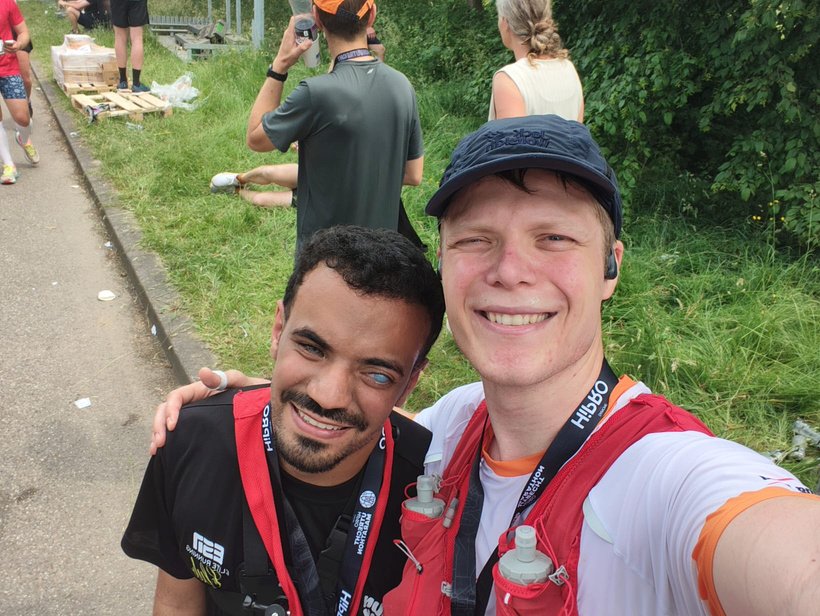 Two runners are smiling together during an event.
In the foreground on the right stands a tall, light‑skinned man wearing a dark blue cap, an earbud in his right ear and a red running vest with two soft water bottles on the front. He is wearing a white T‑shirt with orange trim on the sleeves and has a medal ribbon with text around his neck. He faces the camera with a broad smile and has drops of sweat on his face, as if he has just been running.
Next to him on the left stands a man with a medium to dark skin tone, short black curly hair, and a dark beard and moustache. His right eye is milky white or light blue in colour, his left eye dark; he is smiling kindly toward the camera. He is wearing a black T‑shirt with white and yellow logos on the chest, a black medal ribbon and also a red running vest. The man on the right has his arm around his shoulders.
In the background there is a strip of grass along a road with several people who seem to be taking a break from a run: someone is sitting on the grass with legs stretched out, someone in a dark T‑shirt and orange cap is drinking from a cup, and further away there is a woman with light hair in a ponytail in a light top with her back to the camera. On the left the asphalt of the road is visible, with some cups or litter scattered along the edge. On the right there is dense green shrubbery and trees. The atmosphere feels cheerful and sporty, as if the photo was taken during or after a running event.