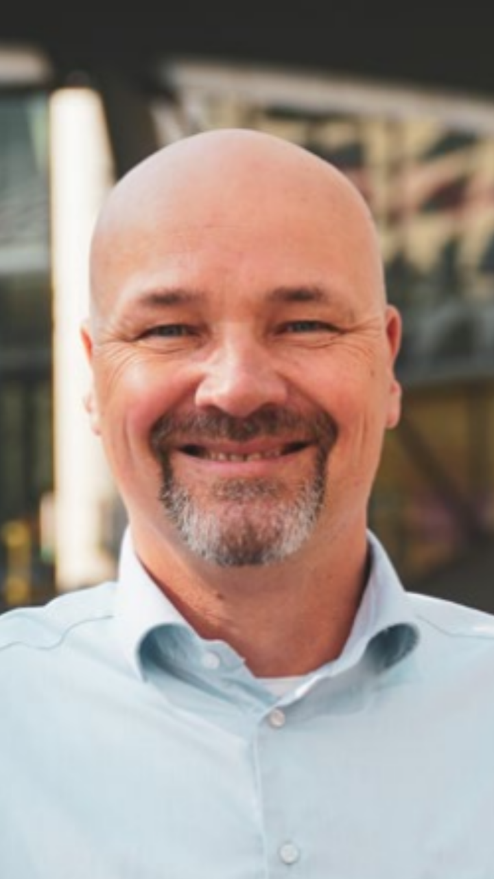 Mark de Buck smiles at the camera. He has a grey goatee and wears a light blue shirt.